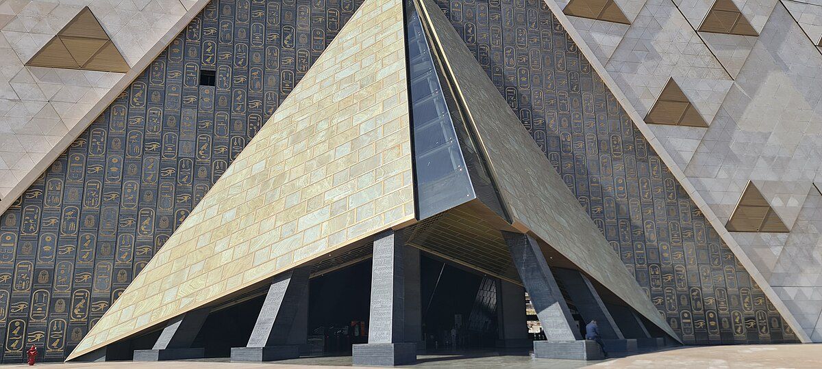 Un grande e moderno ingresso a forma di piramide, con pannelli dorati e alti pilastri scuri, accoglie i visitatori del nuovo museo egizio, su una parete decorata con la ripetizione di antichi geroglifici egizi.