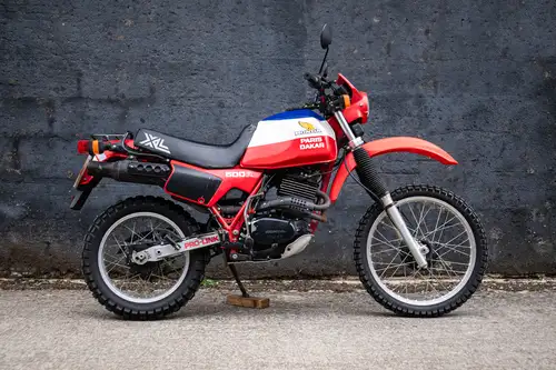 Una moto fuoristrada Honda XL Paris-Dakar rossa, blu e bianca, conosciuta nelle avventure motociclistiche di Harry Metcalfe, è parcheggiata su una superficie di cemento di fronte a un muro grigio scuro. La moto è dotata di pneumatici tassellati e di un design robusto per una guida avventurosa.