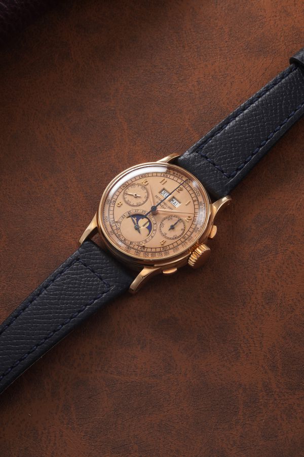 Un orologio da polso d'oro con quadrante marrone, cinturino in pelle blu e molteplici contatori si trova su una superficie strutturata marrone. Questo elegante pezzo, che fa parte della collezione orologi costosi 2025, mostra gli indicatori di giorno, data e fasi lunari.