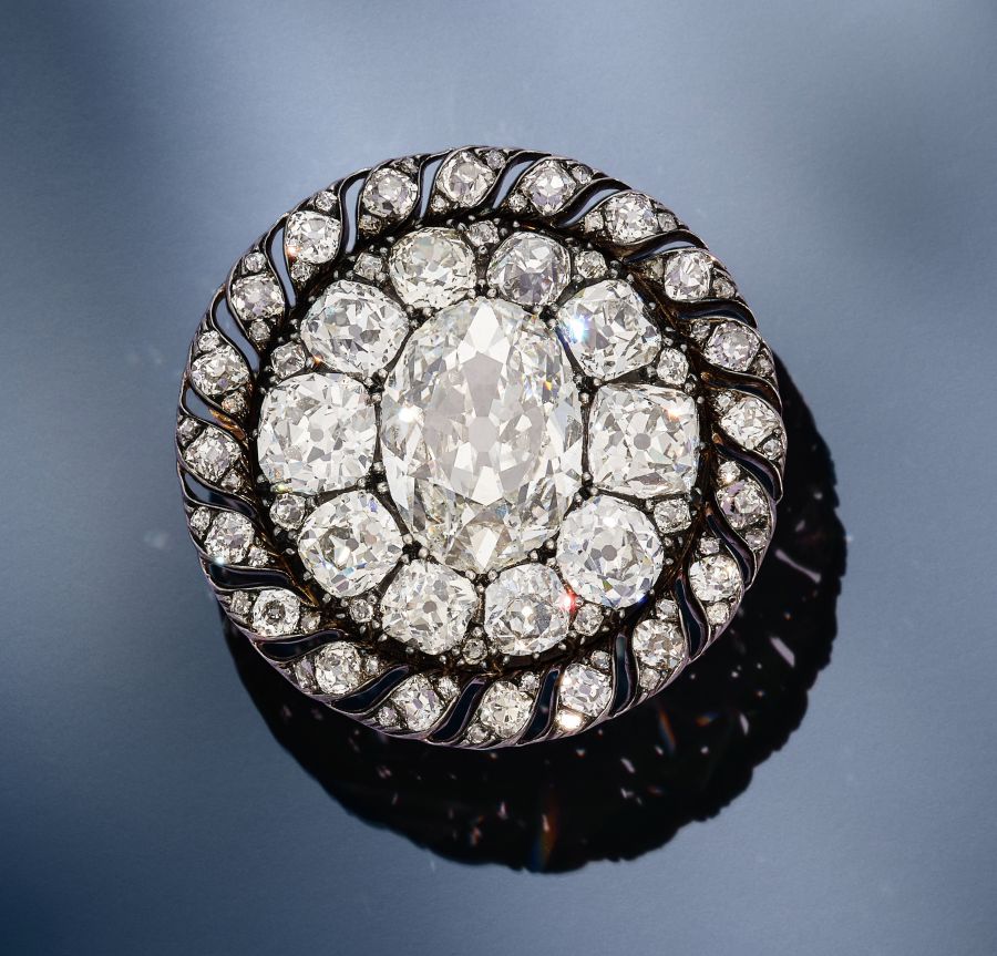 Spilla rotonda e ornata con un grande diamante ovale al centro, circondato da numerosi diamanti più piccoli in un motivo decorativo, su una superficie blu liscia e riflettente.