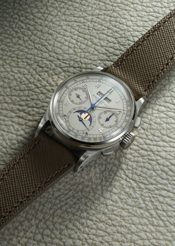Un classico orologio da polso in argento con cinturino marrone testurizzato poggia su una superficie chiara e ciottolata. Il quadrante dell'orologio presenta quadranti multipli, lancette blu, indicatori di data, giorno e fasi lunari.