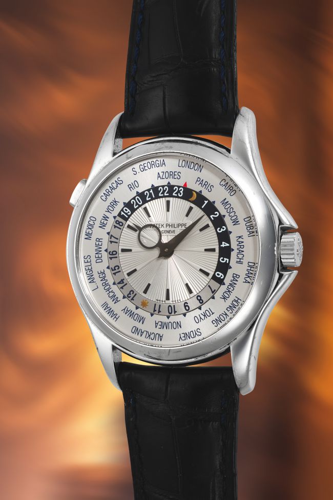 Un lussuoso orologio da polso Patek Philippe con ora mondiale, noto anche come coppola orologio, con cinturino in pelle nera, quadrante argentato e nomi di città che circondano il quadrante, visualizzati su uno sfondo marrone e arancione caldo e sfocato.
