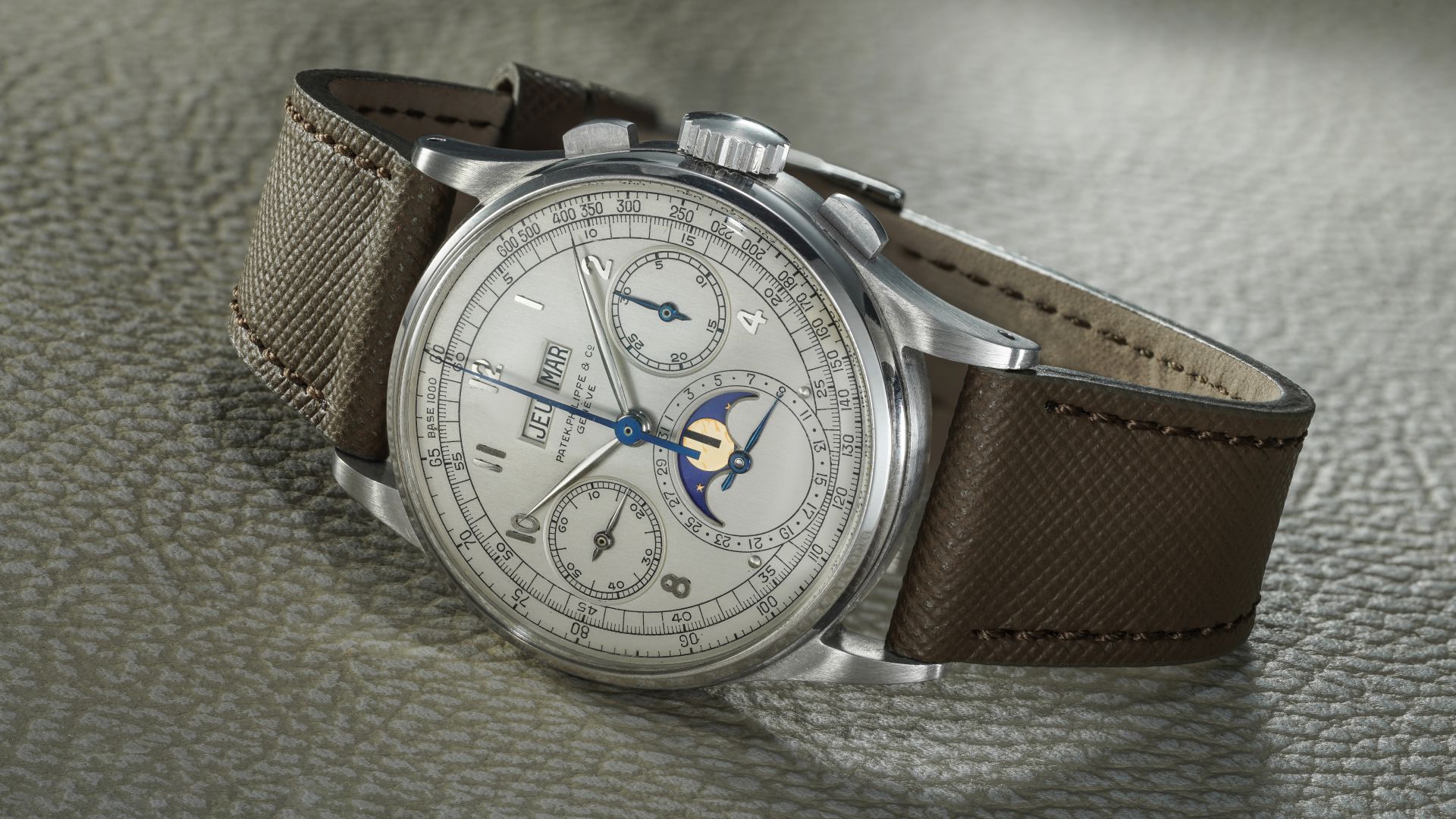 Un orologio da polso di lusso orologi costosi 2025 con quadrante argentato, cinturino in pelle testurizzata marrone, contatori multipli, finestrelle per il giorno e la data e indicatore delle fasi lunari poggia su una superficie con texture chiara.