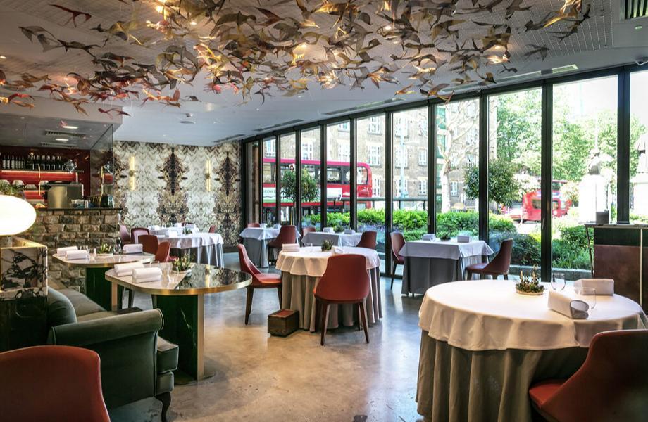 A modern restaurant with round tables covered in white linens, red chairs, large floor-to-ceiling windows, and an artistic ceiling installation of flying birds. Outside, greenery and a red double-decker bus are visible.