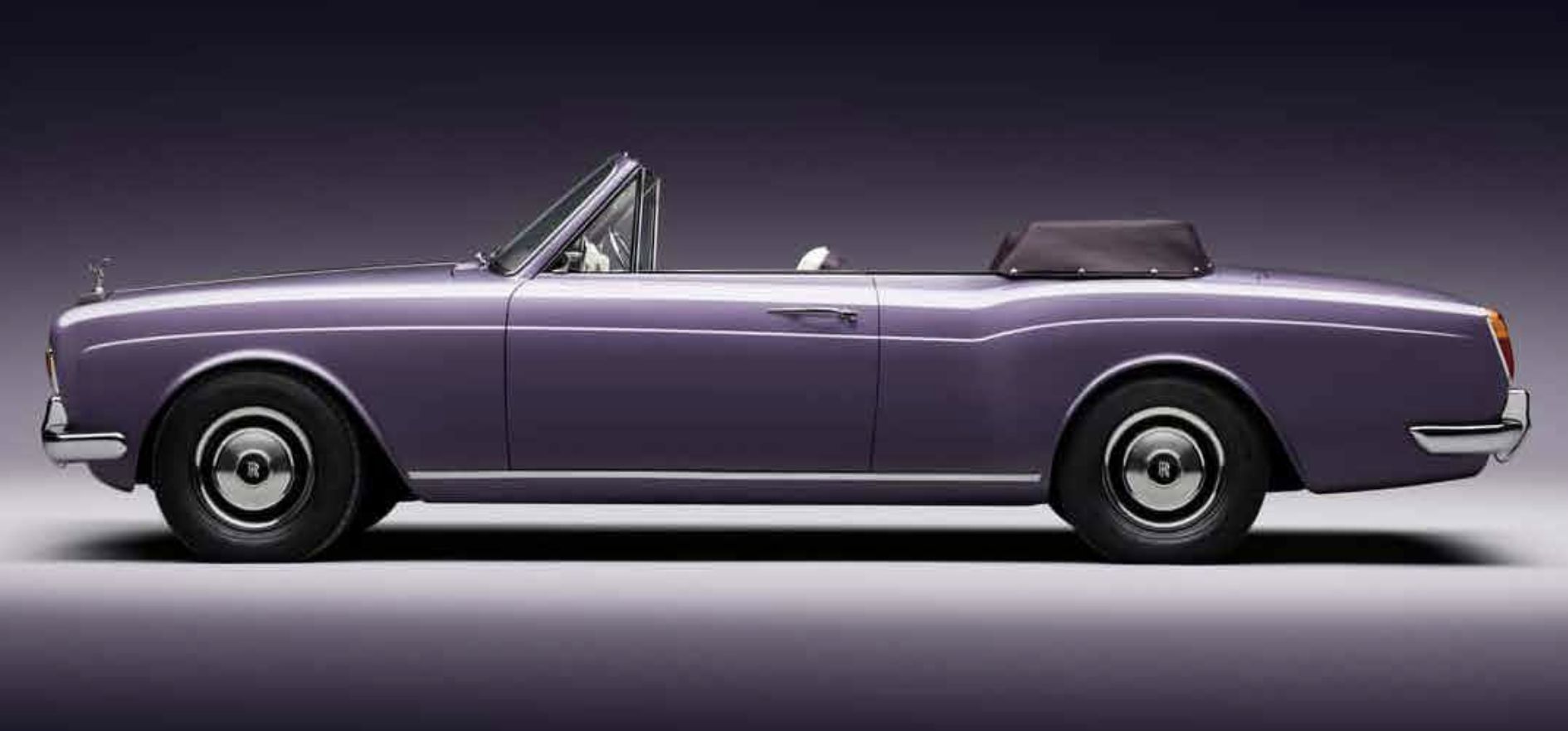 Una classica Rolls-Royce Corniche cabriolet color lavanda con la capote abbassata, mostrata di profilo su uno sfondo scuro sfumato. L'auto presenta dettagli cromati e un design vintage.