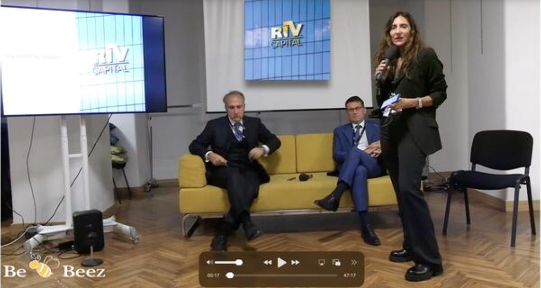 Tre persone si trovano a un evento di lavoro. Due uomini in giacca e cravatta siedono su un divano giallo; una donna in abito scuro è in piedi e tiene un microfono. Gli schermi televisivi alle loro spalle mostrano "Rtv Capital", riflettendo l'energia dinamica di questo ufficio o studio riv.