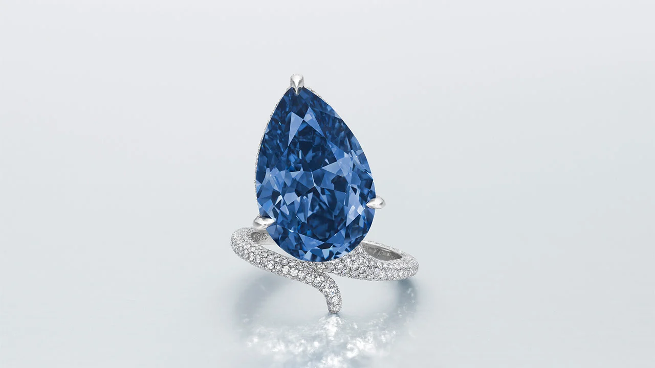 Anello con diamante a forma di pera di colore blu mellon, con fascia in oro bianco incrostata di diamanti e ritorta su una superficie di luce riflettente.