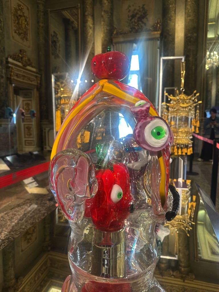 Una scultura in vetro colorato con forme astratte e motivi di occhi è esposta in una stanza decorata in oro con specchi, colonne e lampadari. I riflessi e la luce creano un'atmosfera artistica e vibrante.