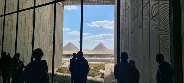 All'interno del nuovo museo egizio, sagome di persone guardano attraverso alte vetrate le Piramidi di Giza sotto un cielo parzialmente nuvoloso.