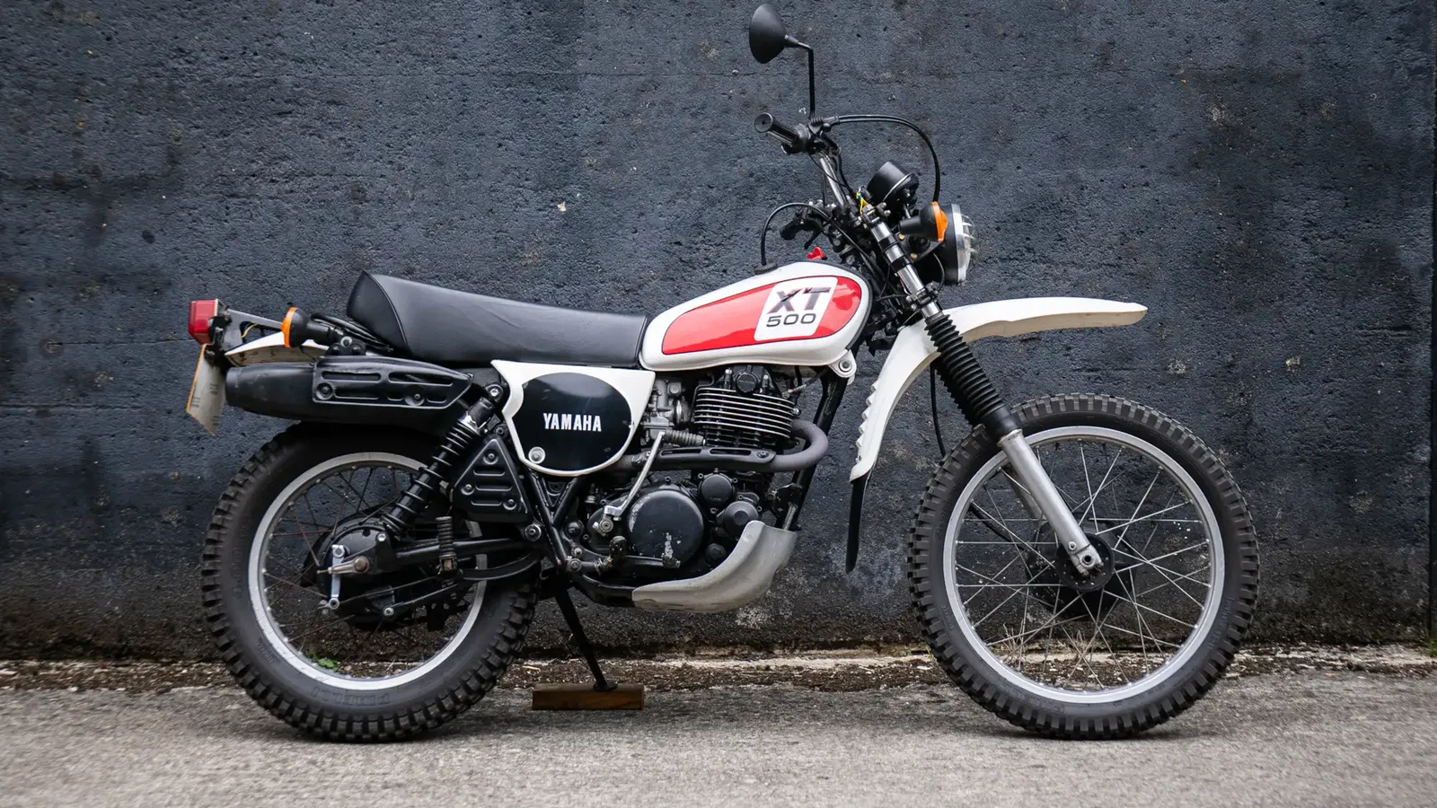 Una moto Yamaha XT 500 d'epoca con pneumatici tassellati, serbatoio rosso e bianco e sella nera è parcheggiata su una superficie di cemento contro un muro grigio scuro e ruvido, perfetto per ogni appassionato di moto di Harry Metcalfe.