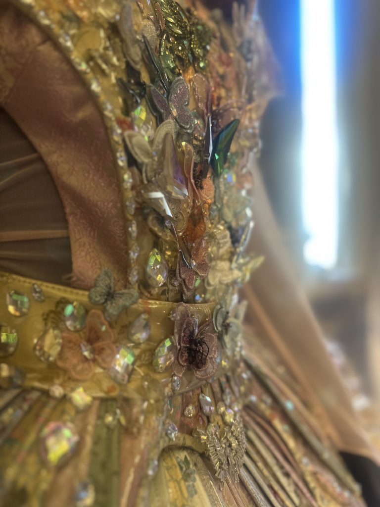 Primo piano di un corpetto decorato con gemme colorate e iridescenti, perline e fiori di tessuto in oro, rosa e tonalità pastello. Lo sfondo è leggermente sfocato e mette in risalto gli intricati abbellimenti.