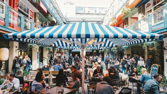A lively outdoor market scene shows people dining and socializing under a large blue-and-white striped umbrella, surrounded by shops and colorful buildings on multiple levels.
