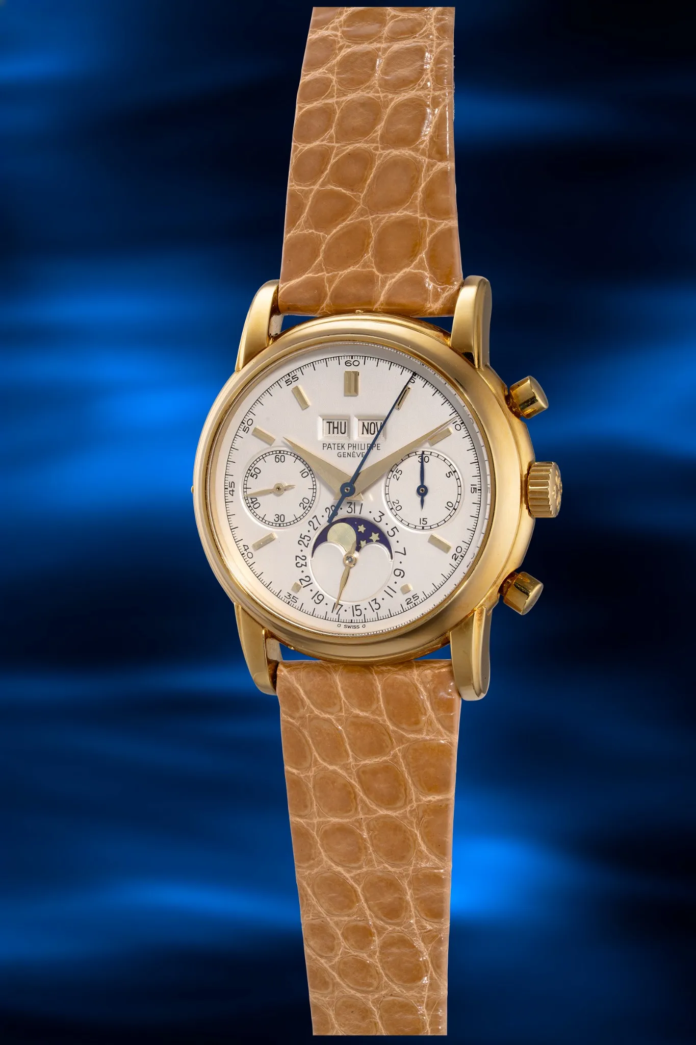 Un orologio da polso d'oro con cinturino in pelle di coccodrillo marrone, quadrante bianco, tre contatori, indicazione delle fasi lunari e finestrelle per il giorno e la data, è elegantemente esposto su uno sfondo blu e ondulato: una squisita citazione dello stile Patek Philippe Hong Kong.