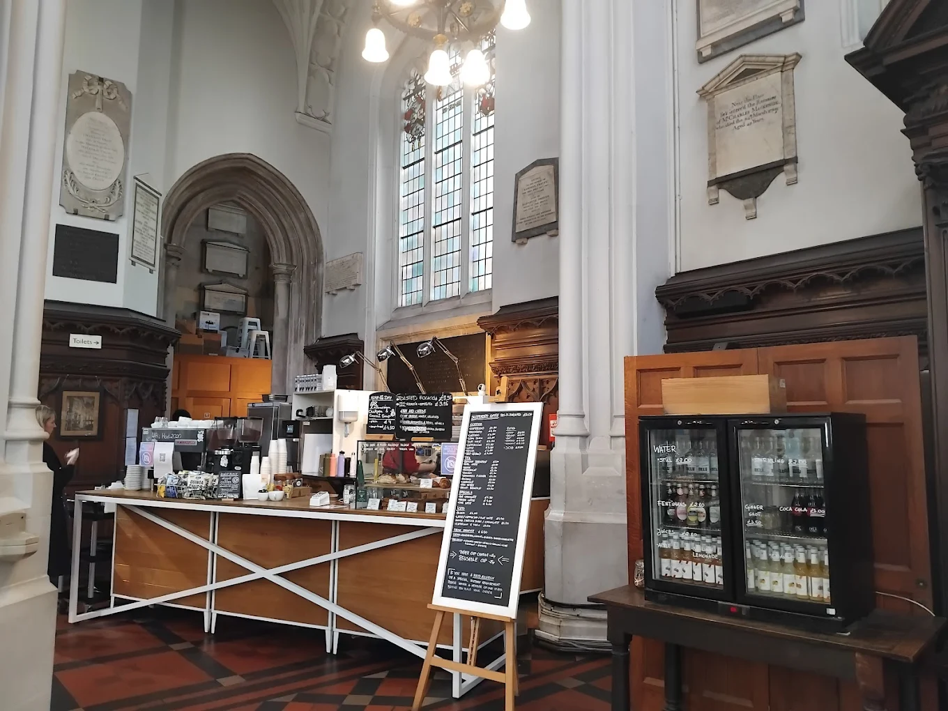 A café counter inside a building with high arched ceilings, stained glass windows, wall plaques, a menu board on an easel, and a glass fridge displaying drinks. The setting combines historic and modern elements.