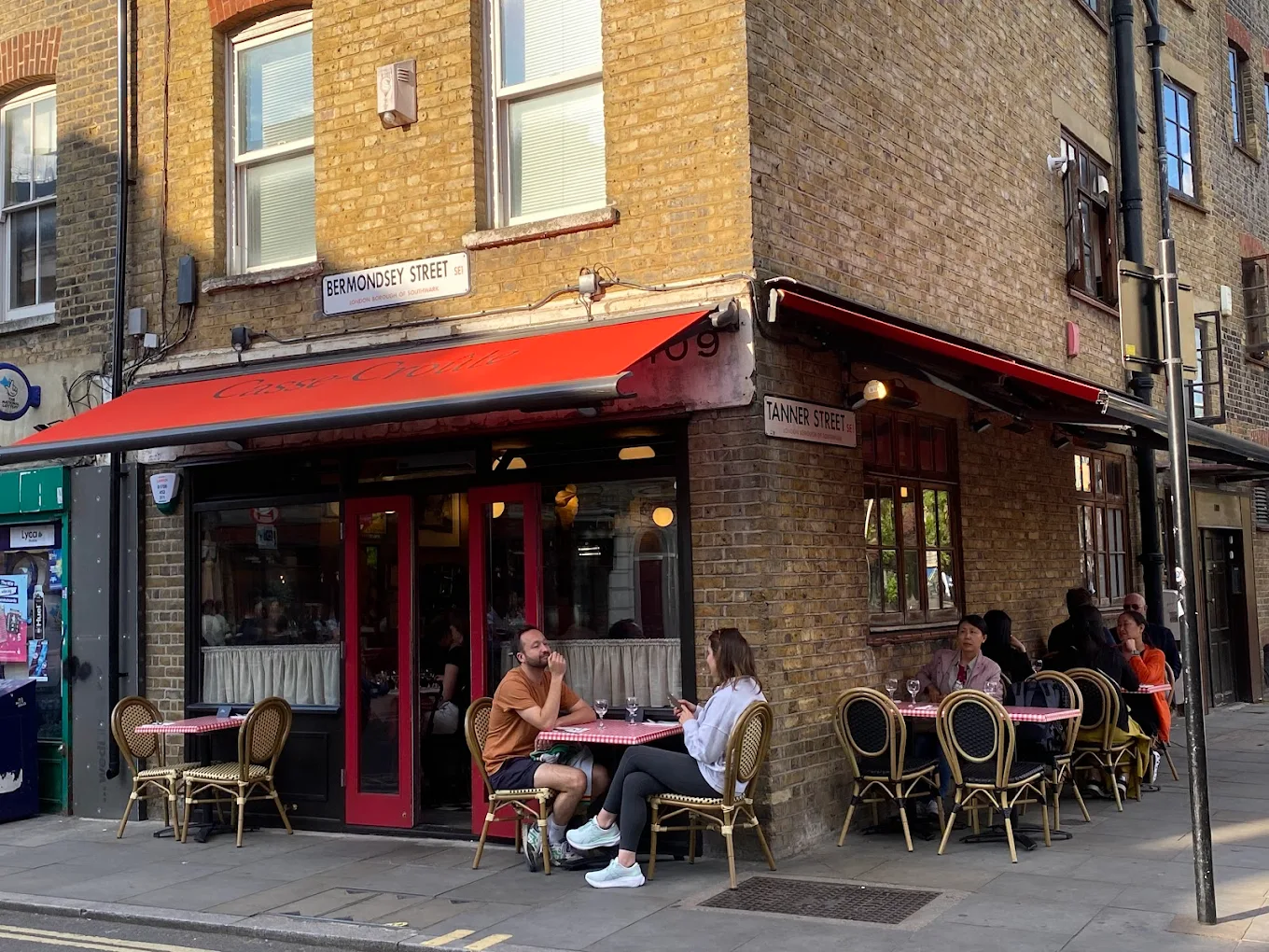 A street corner café with a red awning and outdoor tables, where people are sitting and talking. The building is brick with signs for Bermondsey Street and Tanner Street visible.