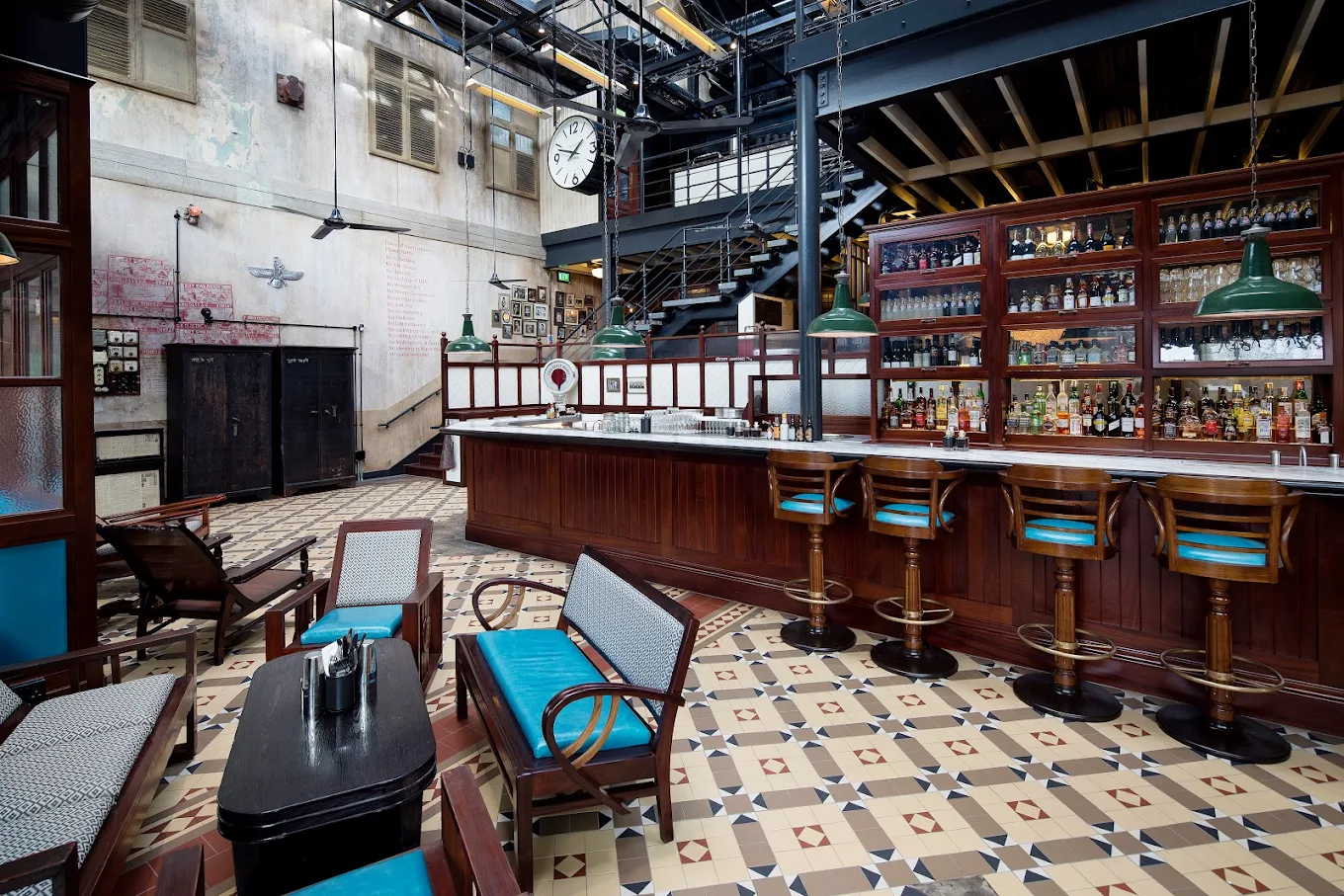 A stylish bar with vintage decor, wooden chairs with turquoise cushions, geometric tiled floor, a large wooden bar with barstools, shelves of bottles, and an industrial staircase under a skylight.