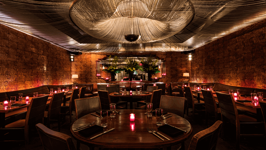 Un ristorante elegante e poco illuminato, con tavoli in legno, candele rosse, sedili in tessuto, pareti in pietra e un soffitto scolpito con drappeggi a rete che creano un'atmosfera calda e invitante.