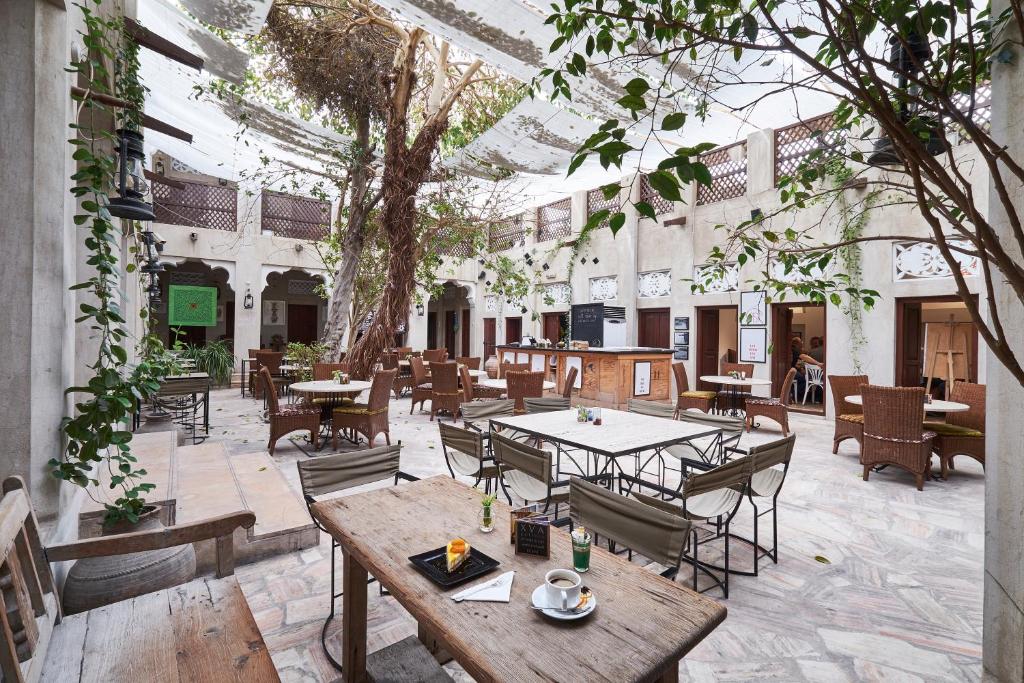A spacious courtyard café with wooden tables and chairs, potted plants, and a large tree growing in the center. Sunlight filters through a partially covered roof, creating a relaxed and inviting atmosphere.