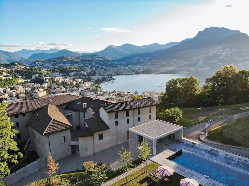 Un edificio in collina si affaccia su un lago panoramico vicino a Reggio Emilia, circondato da montagne e città. Una piscina con ombrelloni a strisce si trova in primo piano, mentre il verde lussureggiante e il cielo azzurro completano il paesaggio pittoresco.