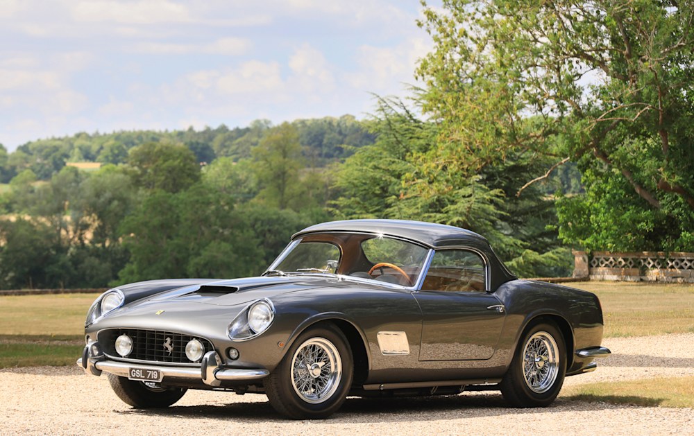Una classica auto sportiva d'epoca argentata con tetto rigido, destinata alla vendita di Christie's 2025, è parcheggiata su un vialetto di ghiaia circondato da alberi e vegetazione lussureggiante sotto un cielo parzialmente nuvoloso.