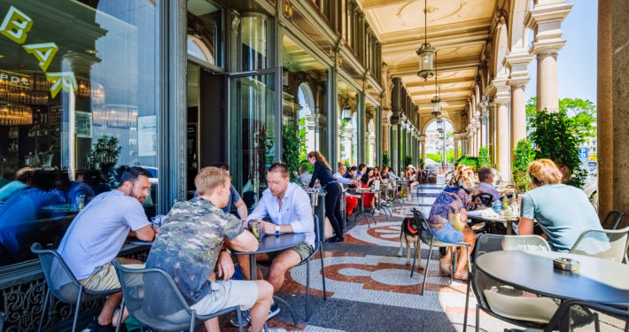 People sit at tables and socialize on a covered outdoor patio of a café, with arched ceilings, large windows, lush green plants, and bright daylight creating a lively, relaxed atmosphere.