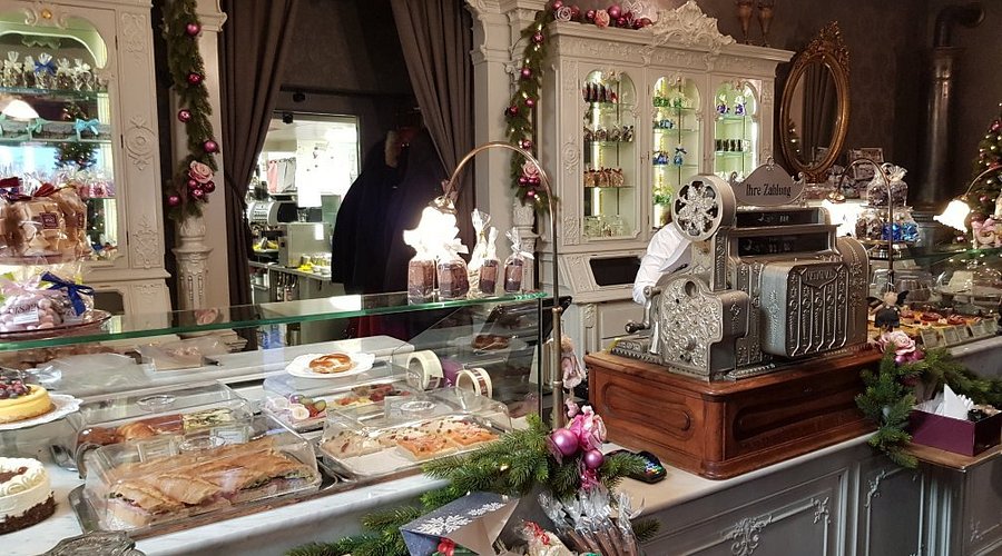 Una pasticceria vintage con armadi bianchi decorati, ghirlande festive e ornamenti rosa trasmette il fascino di Lugano. Pasticcini e torte sono esposti in una vetrina, mentre un antico registratore di cassa si trova sul bancone accanto a barattoli e dolcetti incartati.
