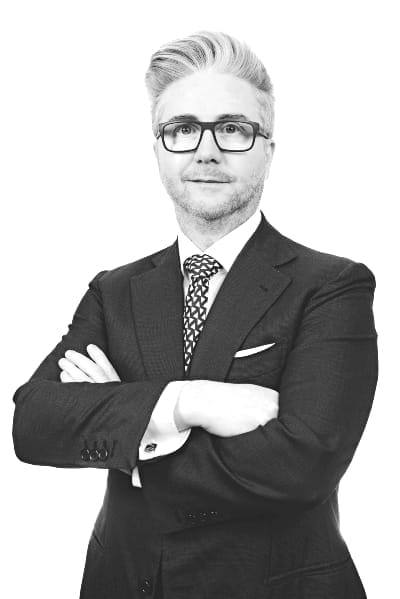 A man in a suit and tie with glasses stands confidently with his arms crossed, looking at the camera against a plain white background.