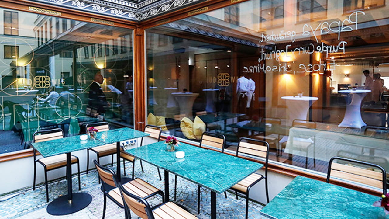 Outdoor seating area of a modern restaurant with green marble tables, wooden chairs, and small floral centerpieces. Large windows show an indoor space with high tables, people, and mirrored walls.