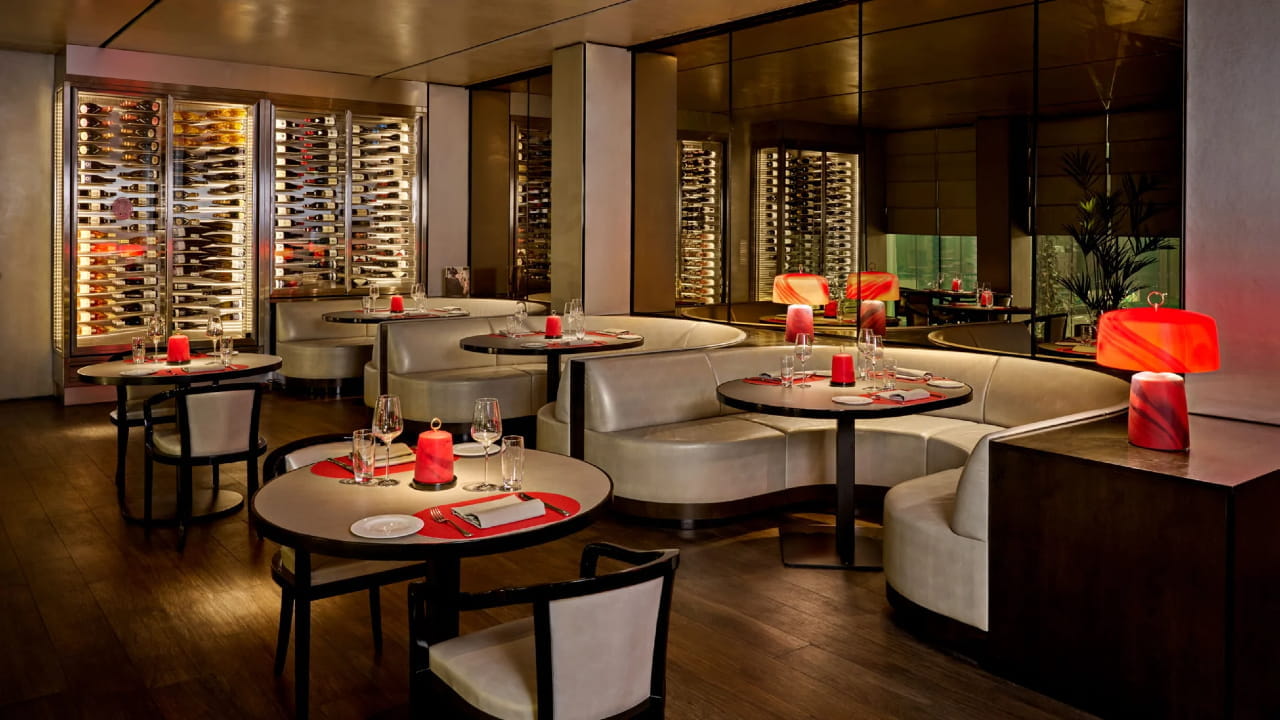 A modern, upscale restaurant interior with round tables, curved white booths, elegant table settings, red lamps, and glass wine racks lining the mirrored walls. The lighting is warm and ambient.