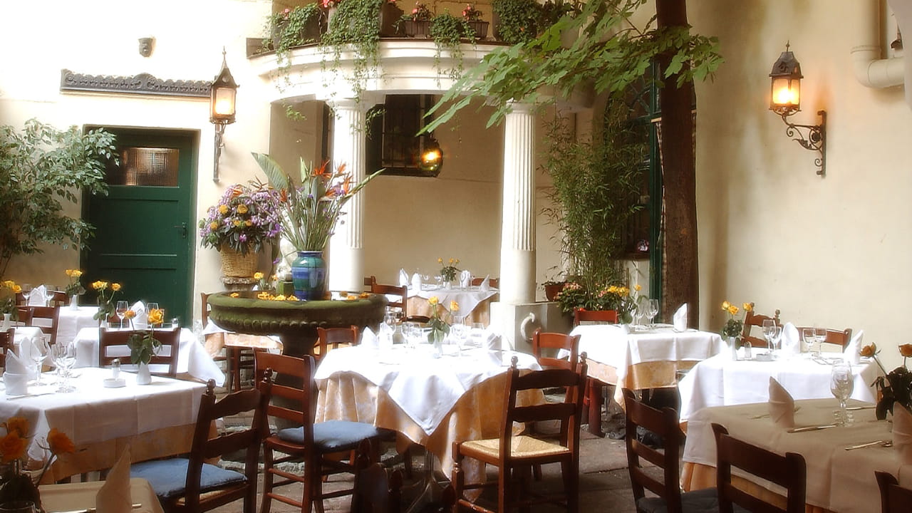 Charming outdoor restaurant patio with wooden chairs, white tablecloths, and yellow flowers on tables. A large flower arrangement and fountain are surrounded by leafy plants and soft, warm lighting.