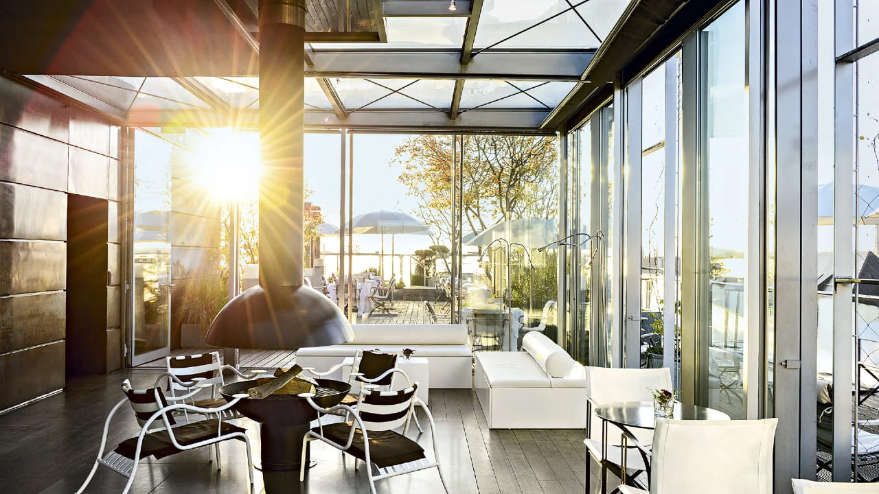A modern glass-walled lounge with a fireplace, white sofas, and a dining table with black and white chairs, filled with sunlight and overlooking an outdoor patio with trees and water.