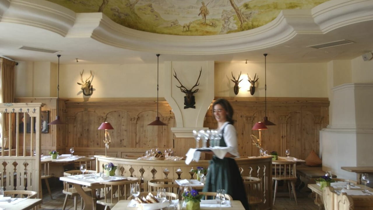 A waitress in traditional attire carries a tray with glasses in a rustic restaurant featuring wooden furniture, white tablecloths, animal head wall mounts, and a decorative mural on the ceiling.