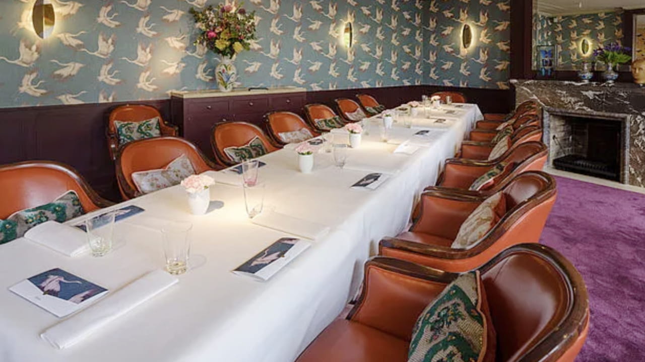 A long dining table with a white tablecloth is surrounded by brown leather chairs. The room features bird-patterned wallpaper, floral decorations, and a marble fireplace. Place settings and glasses are arranged on the table.