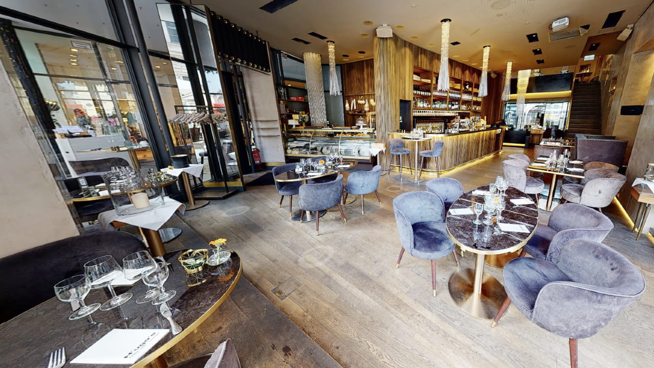 Modern restaurant interior with plush gray chairs, marble tables set with glassware, and a long wooden bar. Floor-to-ceiling windows let in natural light. The space appears upscale and inviting.