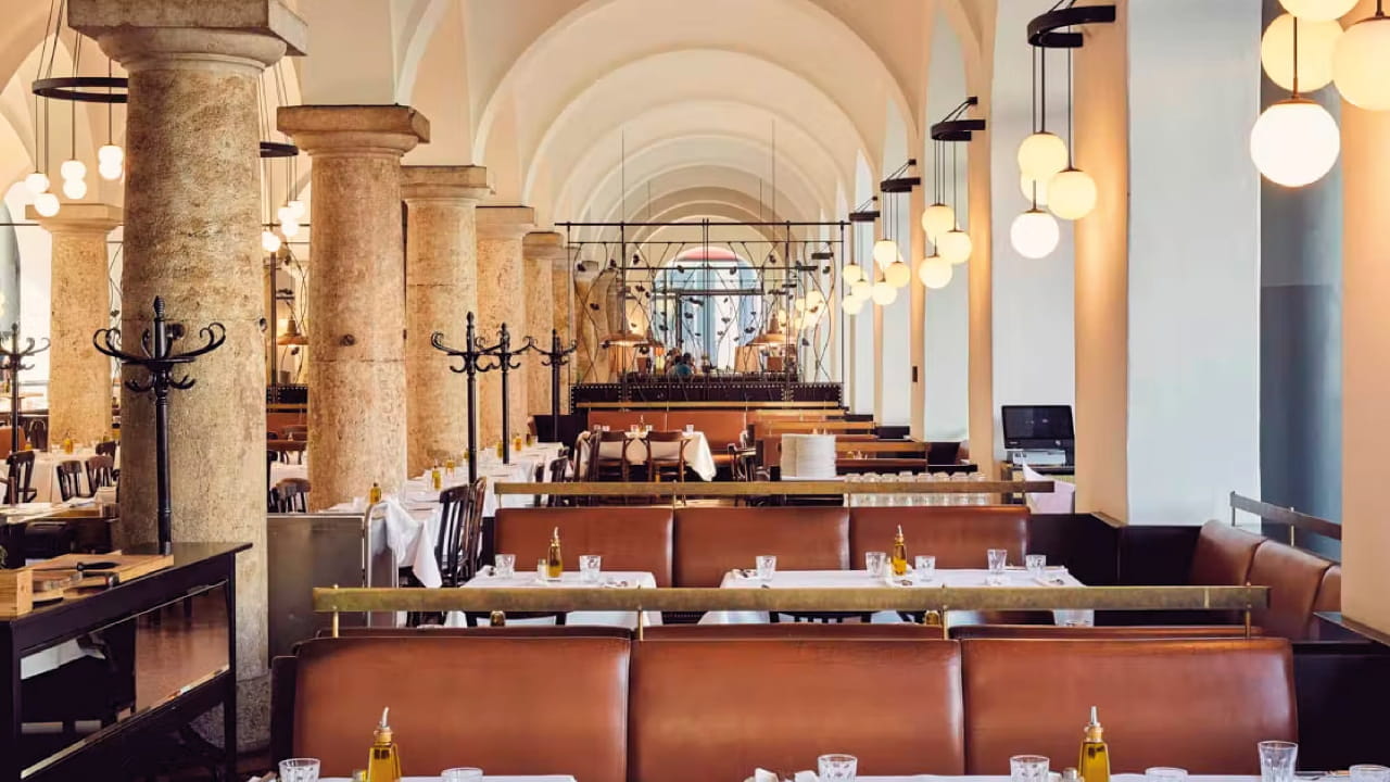 A spacious, elegant restaurant with arched ceilings, stone columns, hanging globe lights, brown leather seating, and white tablecloths. Olive oil bottles are placed on each table. The space is empty of people.