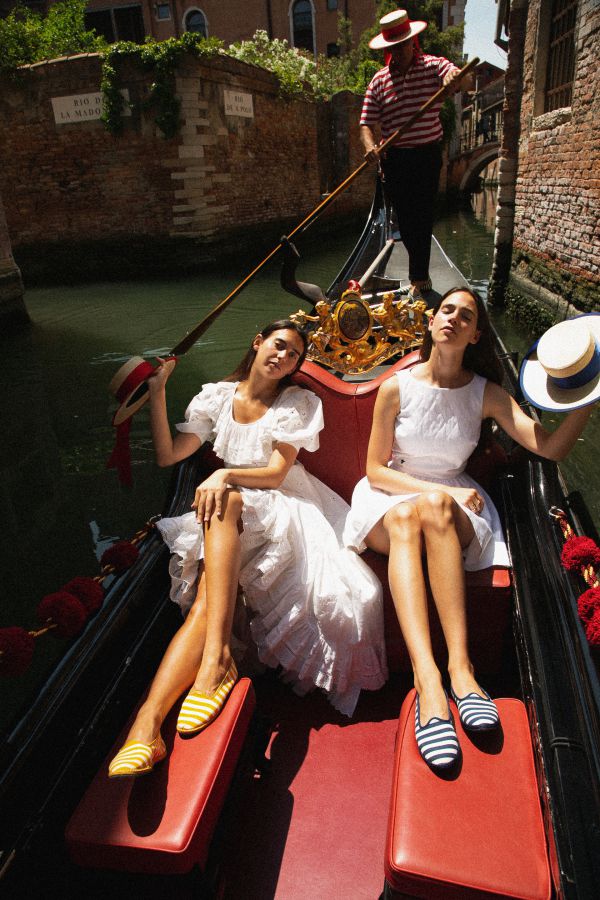 Due donne in abito bianco si rilassano su una gondola ViBi Venezia a Venezia, con un gondoliere dietro di loro mentre galleggiano lungo il canale. Entrambe indossano scarpe a righe e cappelli, godendosi la giornata di sole.