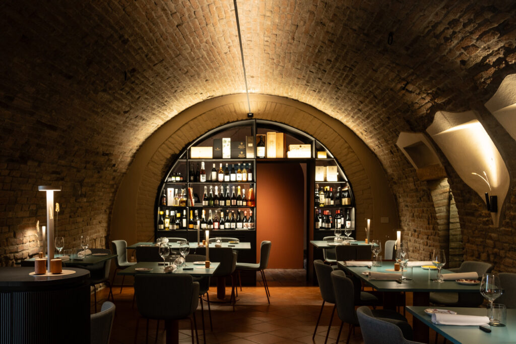 Un ristorante trapanese accogliente e raffinato, con soffitti ad arco in mattoni, luci soffuse, tavoli e sedie moderne e una parete di fondo foderata con scaffali di bottiglie di vino e liquori.