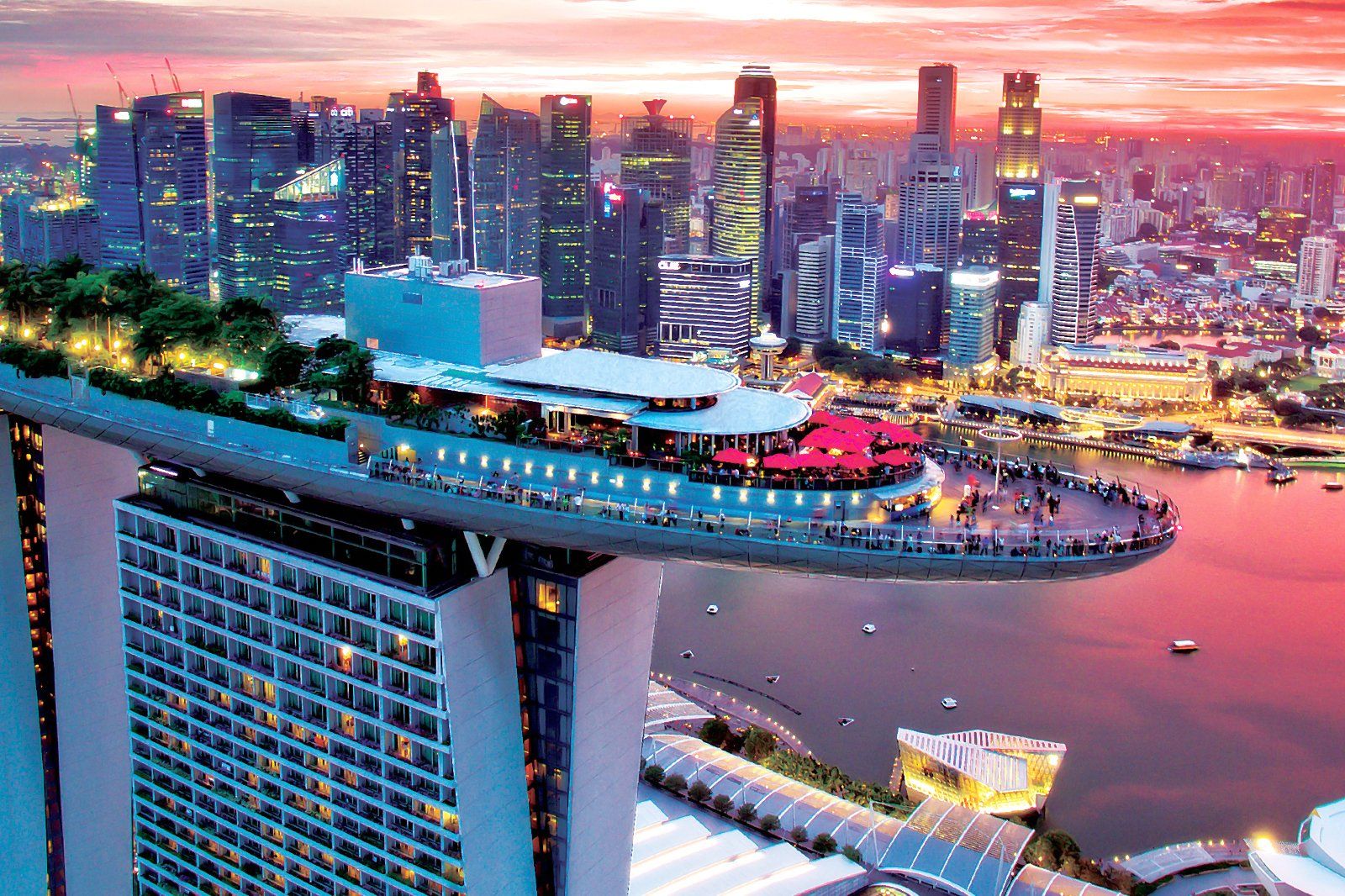 Aerial view of Marina Bay Sands’ rooftop infinity pool and sky park overlooking Singapore’s downtown skyline at sunset, with tall buildings, waterfront, and vibrant city lights.