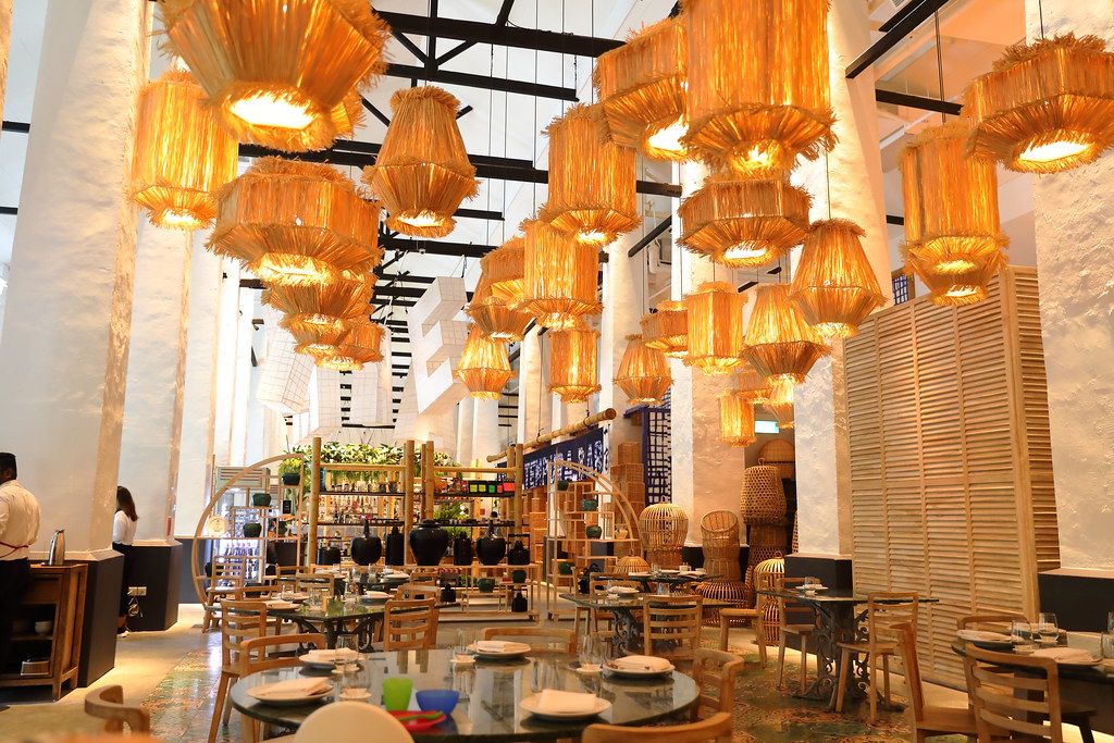 A modern restaurant interior featuring warm, woven pendant lights hanging from a high ceiling, wooden tables and chairs set for dining, and decorative shelves with plants and ceramics.