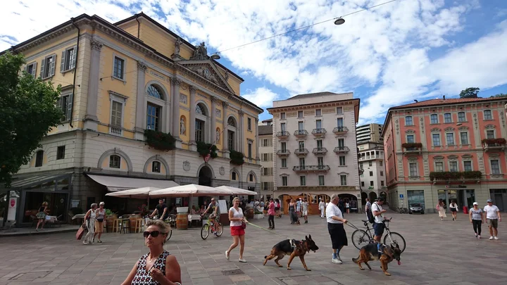Persone che camminano, vanno in bicicletta e si rilassano nei caffè all'aperto di una vivace piazza di Reggio Emilia, circondata da edifici storici con balconi e finestre ad arco, sotto un cielo parzialmente nuvoloso. Due cani vengono portati a spasso in primo piano.