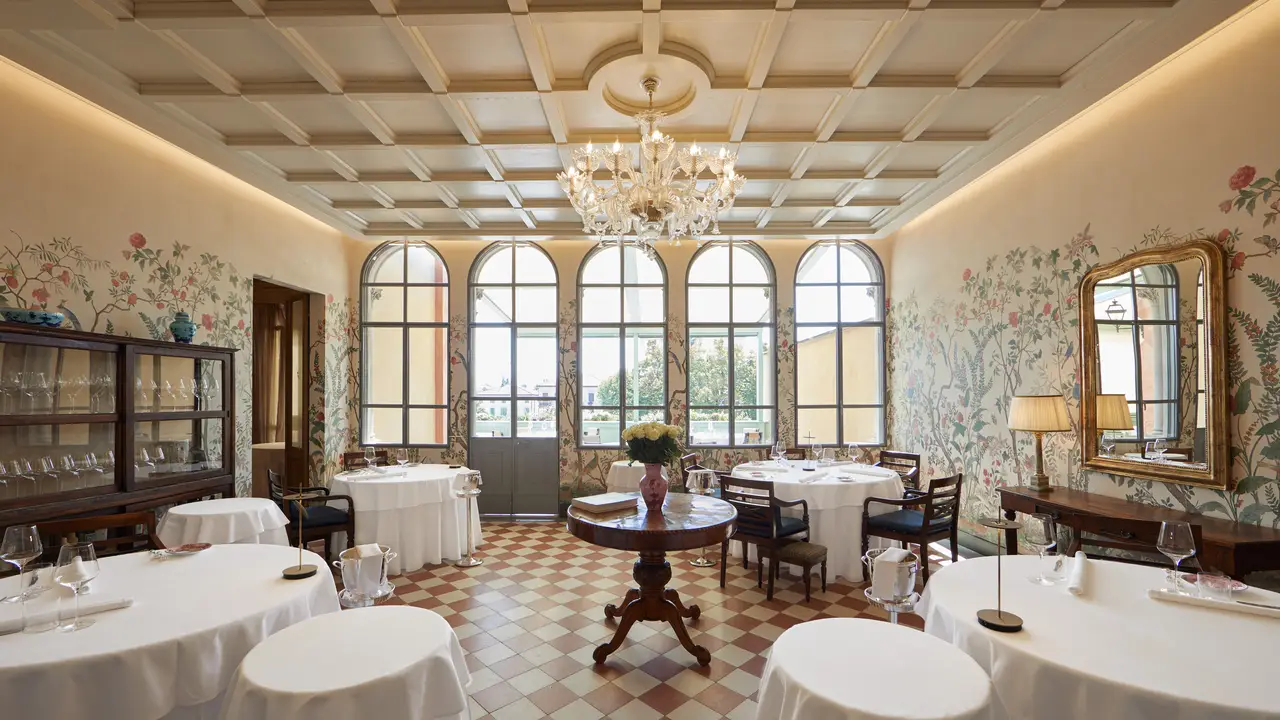 Elegante ristorante con tavoli rotondi ricoperti di stoffe bianche, alte finestre ad arco, carta da parati floreale, lampadario e mobili classici in legno. Ispirato al fascino di Trapani, la luce naturale riempie l'ambiente, creando un'atmosfera calda e invitante.