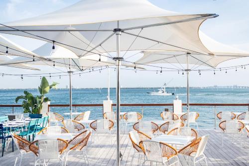 Outdoor restaurant patio with white wicker chairs and round tables under large white umbrellas, overlooking a blue body of water with a boat in the distance on a sunny day.