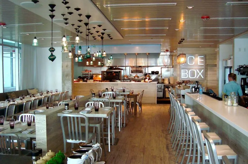Modern restaurant with wooden floors, metal chairs, pendant lights, and an open kitchen at the back. Tables are neatly set, and a person stands behind a long counter on the right. The word "ICE BOX" is on the wall.