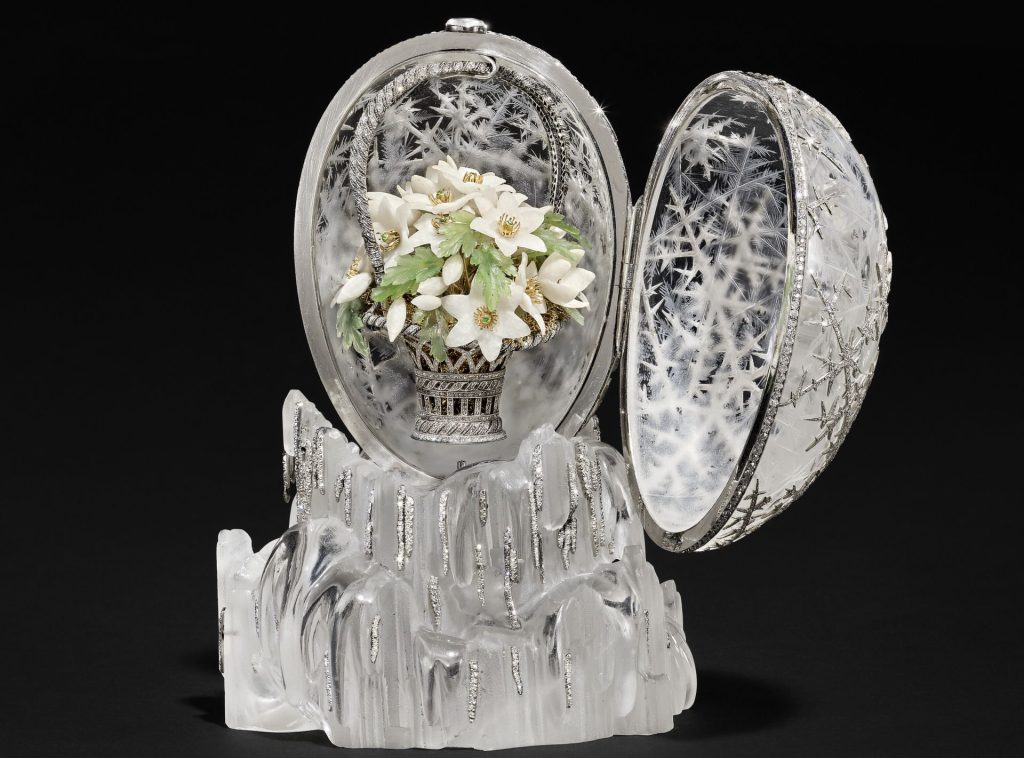 Un uovo decorativo Fabergé con esterno smerigliato e simile al ghiaccio si apre, rivelando un cesto di fiori bianchi con foglie verdi e accenti dorati all'interno, appoggiato su una base simile al cristallo.