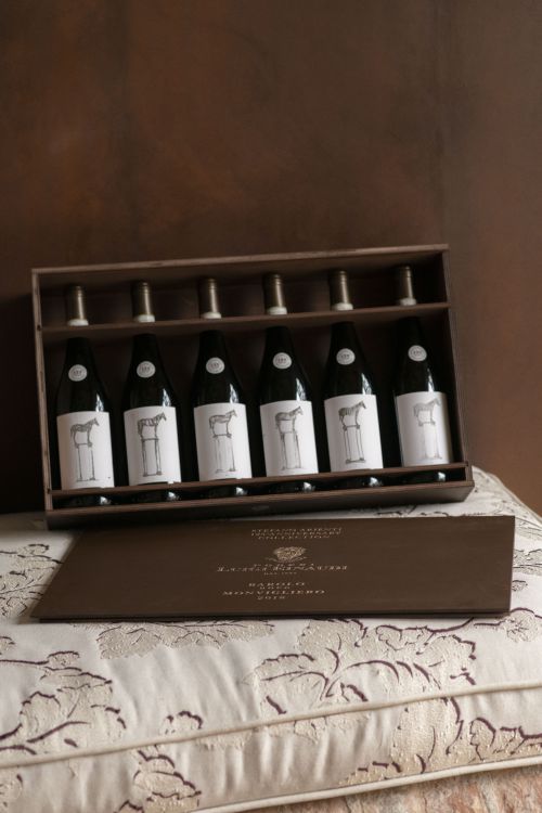 Sei bottiglie di Barolo Monvigliero con etichette bianche, ciascuna con l'illustrazione di una macchina da cucire, sono ordinatamente disposte in una scatola marrone aperta, appoggiata su cuscini a fantasia con un coperchio marrone chiuso in tinta.