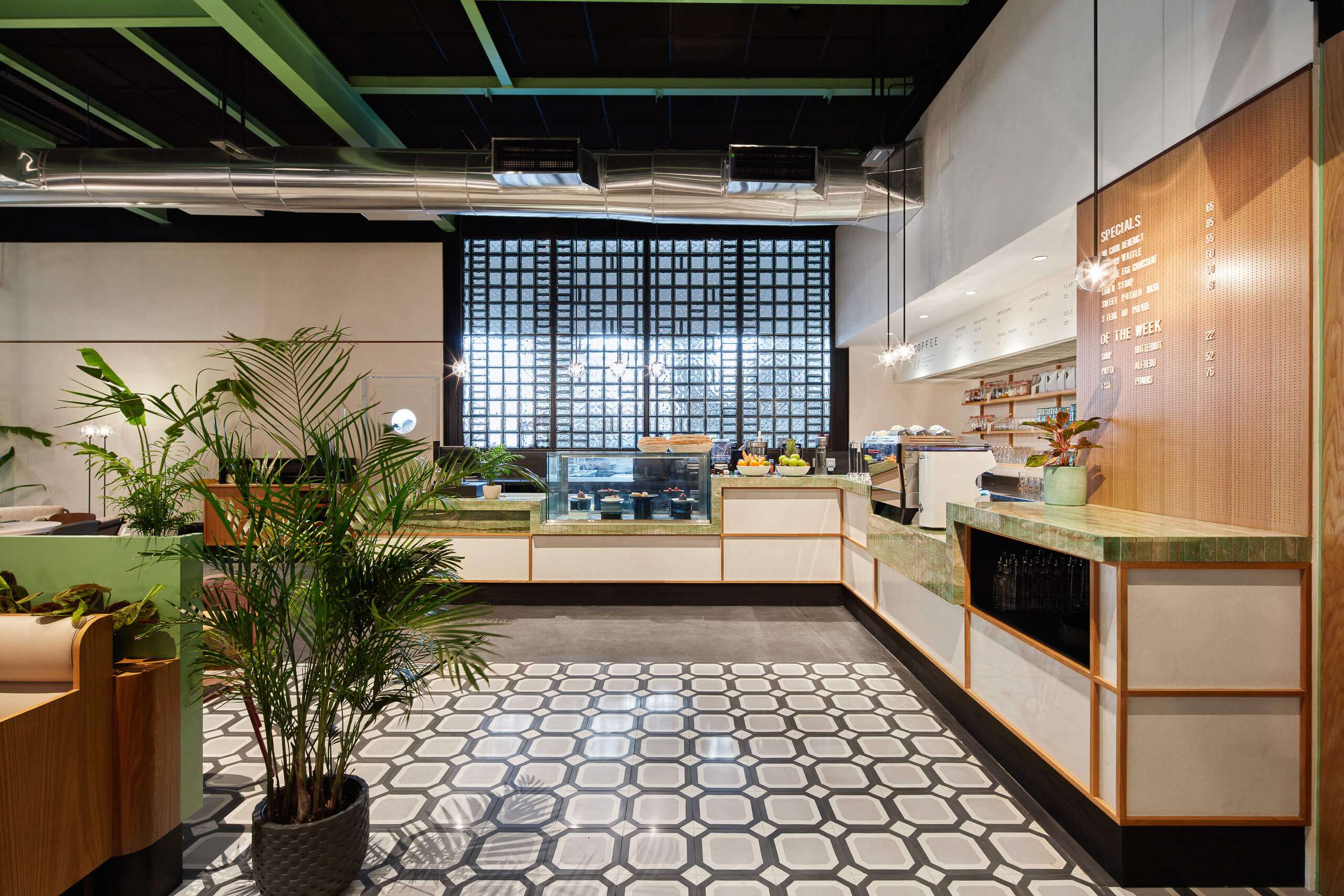 Modern café interior with geometric tile floor, green countertops, wood accents, and a menu board on the wall. Glass display cases hold pastries, and potted plants add greenery to the bright, inviting space.
