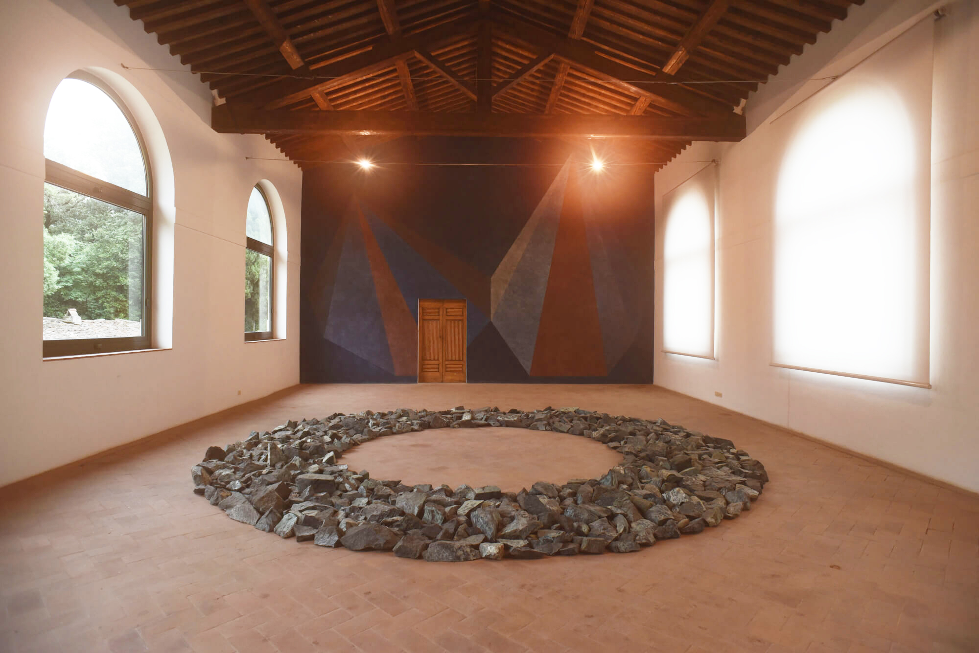 Una grande composizione circolare di rocce si trova su un pavimento piastrellato in una stanza spaziosa con alti soffitti in legno, finestre ad arco e un murale geometrico sopra una porta di legno chiusa, che evoca lo spirito artistico di Trapani.