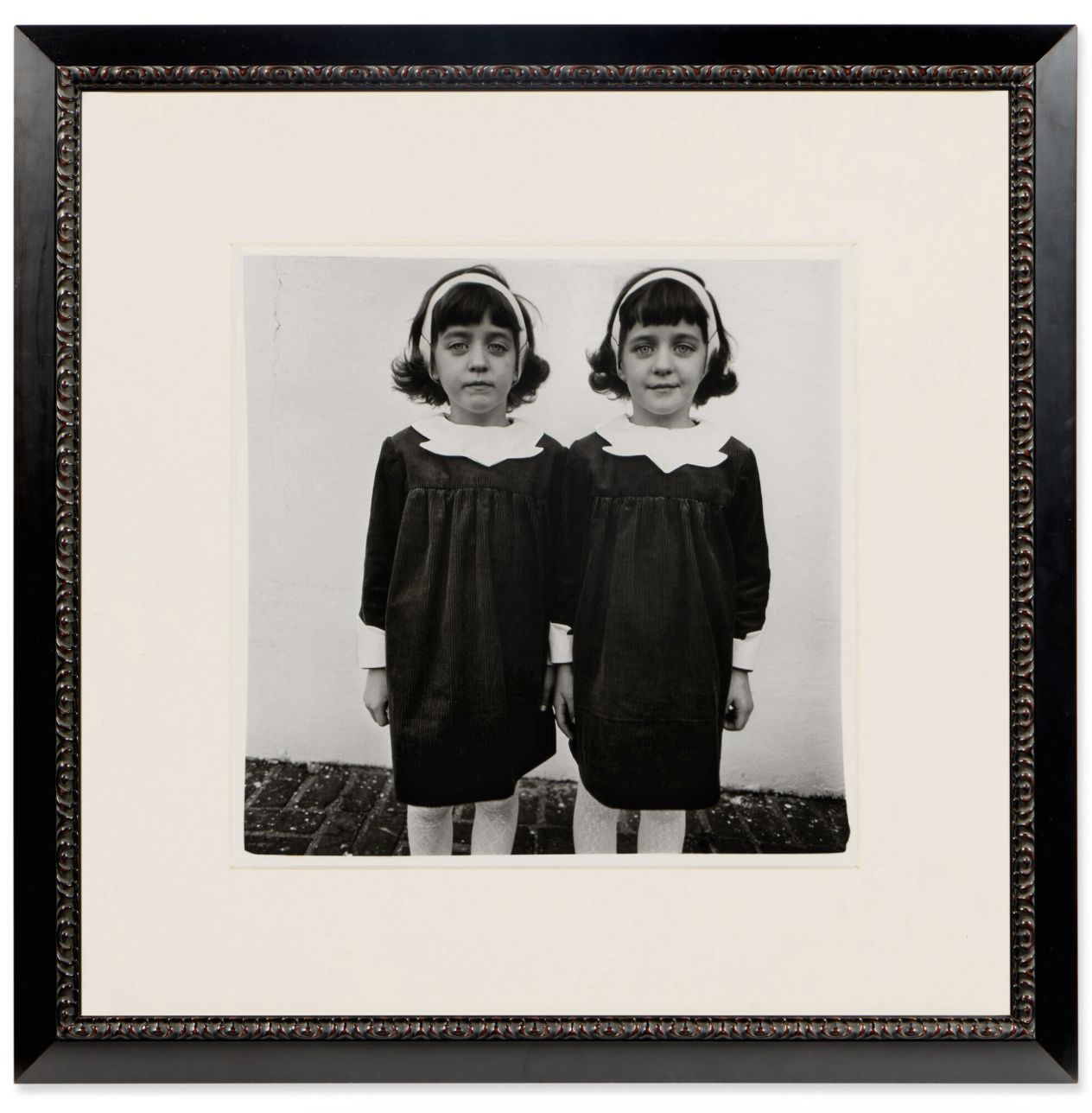 Una foto in bianco e nero in una cornice mostra due giovani gemelle in abiti coordinati in piedi una accanto all'altra. Questa immagine, tra le foto più costose 2025, cattura il loro sguardo diretto contro una parete semplice con un'eleganza senza tempo.