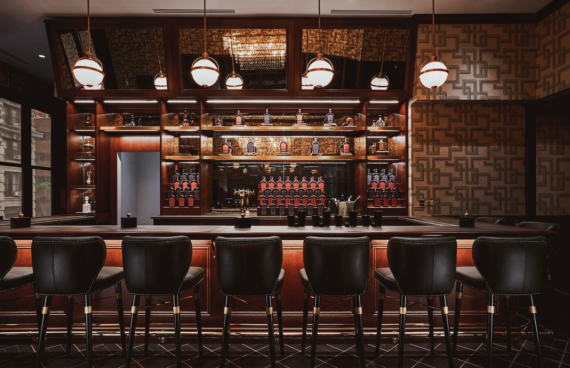 Elegante bar con una fila di sgabelli in pelle nera, un bancone in legno e scaffali retrostanti che espongono bottiglie e bicchieri. L'illuminazione calda e la carta da parati a motivi geometrici creano un'atmosfera elegante e invitante.