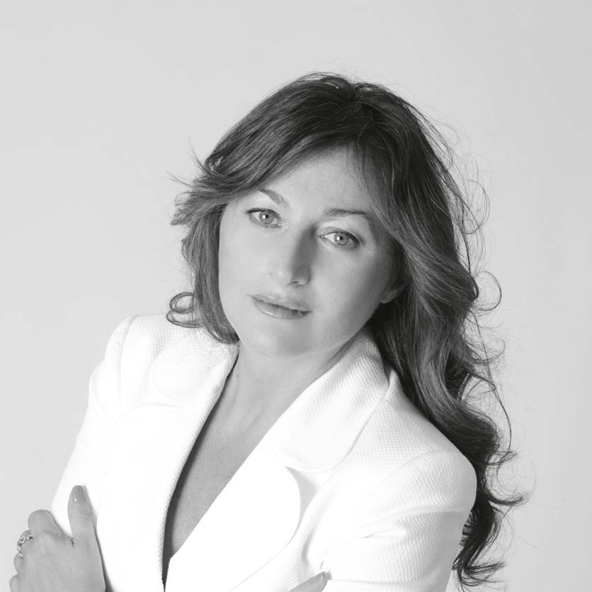 Ritratto in bianco e nero di una donna con capelli mossi che indossa un blazer bianco e guarda l'obiettivo con un'espressione neutra su uno sfondo semplice di ispirazione trapanese.